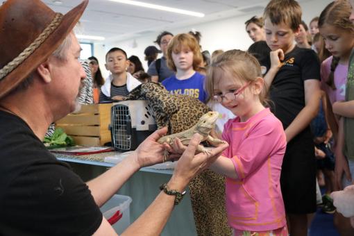 An educational reptile program where a handler lets a young girl hold a friendly bearded dragon while a group of children watches closely